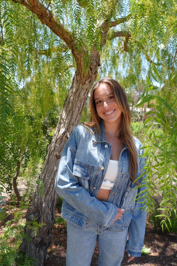 Young woman with long hair smiling outdoors, standing in front of a tree with green foliage.