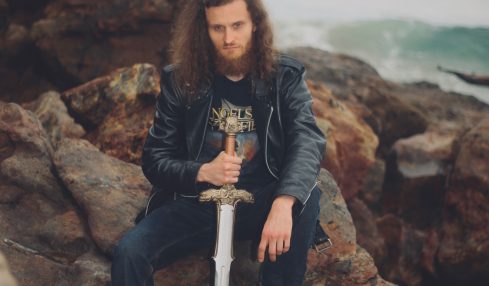 Person with long curly hair holding a sword, sitting on rocks near the ocean, wearing a leather jacket and jeans.