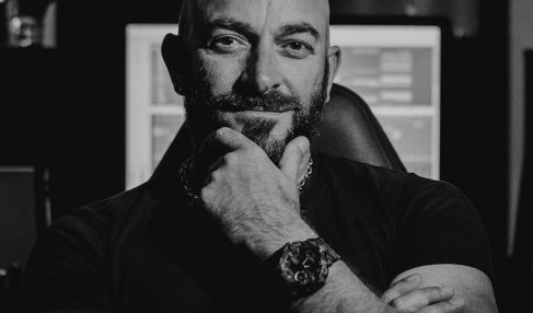 Man with a beard and tattooed arm, sitting with hand on chin, in a dark room with computer screens behind him.