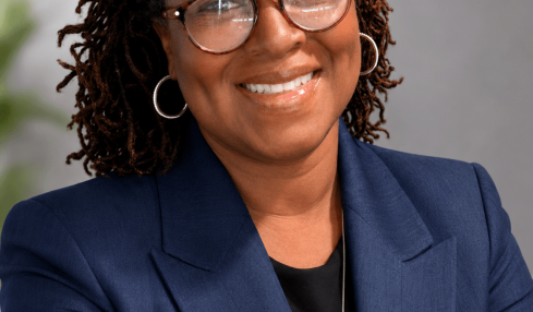 Portrait of a smiling woman with glasses, wearing a navy blazer and black top, against a neutral background.