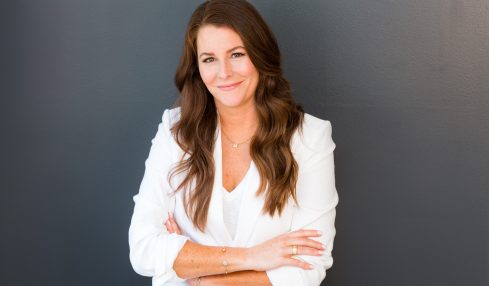 Woman with long brown hair smiling, arms crossed, wearing a white blazer, standing against a dark background.
