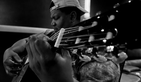 Person playing guitar, wearing a cap backwards, in a dark setting, focused on the instrument.