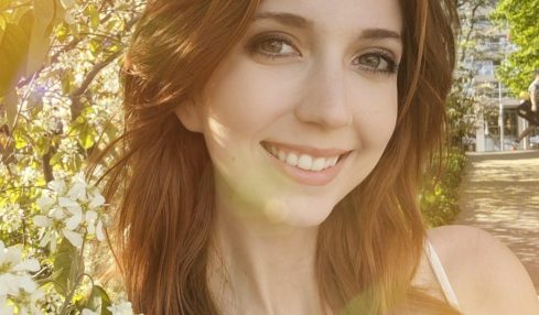 Young woman smiling outdoors with blooming flowers and sunlight, surrounded by green leaves and trees.