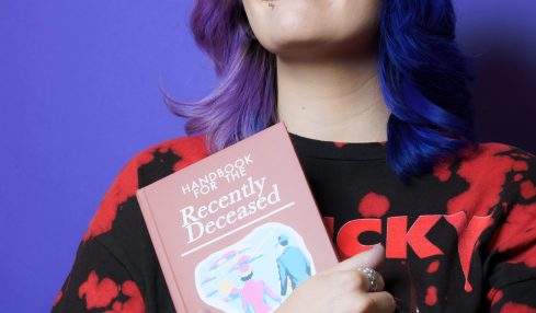 Woman with colorful hair holding a book, smiling, against a purple background.