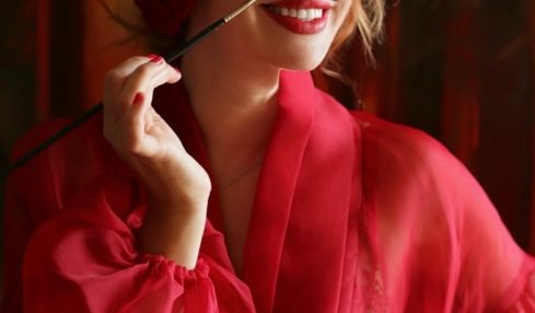 Woman with red hair and lipstick, wearing a red silk robe, smiling and holding a cigarette holder.