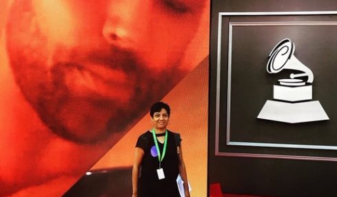 Person standing on red carpet in front of large screen with a record player icon, large face image in background.