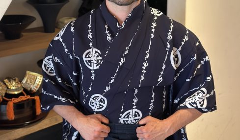 Man in traditional Japanese attire standing indoors with shelves of bowls behind him.