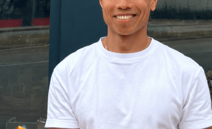 Smiling man in white t-shirt and blue cap holding a tray of assorted donuts outdoors.