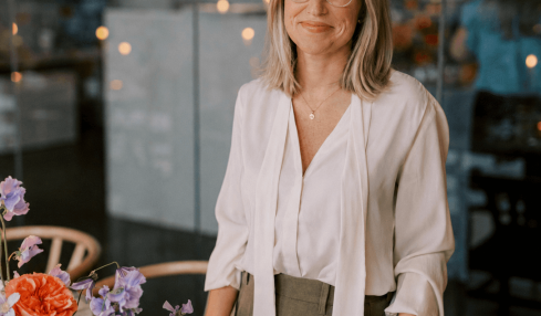 Woman with glasses and shoulder-length hair stands near a table with colorful flowers in a modern indoor setting.