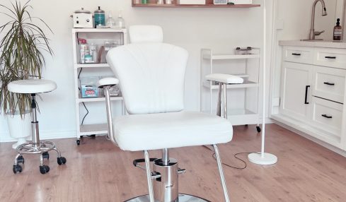 Medical or beauty treatment room with a white chair, shelves, and a lamp, on a wooden floor.