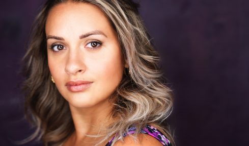Portrait of a woman with wavy blonde hair, wearing a colorful top, against a dark background.