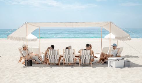 People relaxing under a canopy on a sandy beach with the ocean and sky in the background.