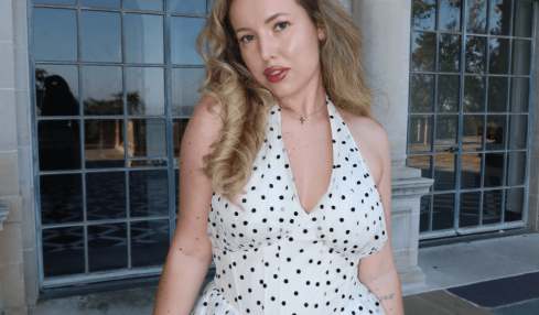 Young woman with wavy hair in a polka dot dress standing outside a building with large arched windows.