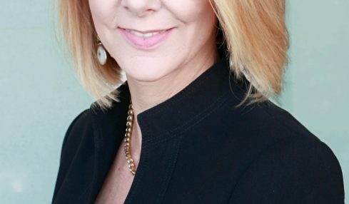 Woman with blonde hair and glasses smiling, wearing a black blazer, against a light background.