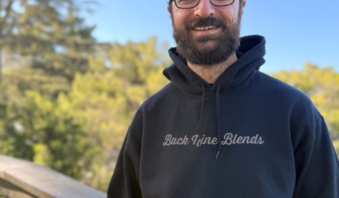 Smiling man with glasses, beard, black hoodie, and backwards cap outdoors with trees and blue sky.