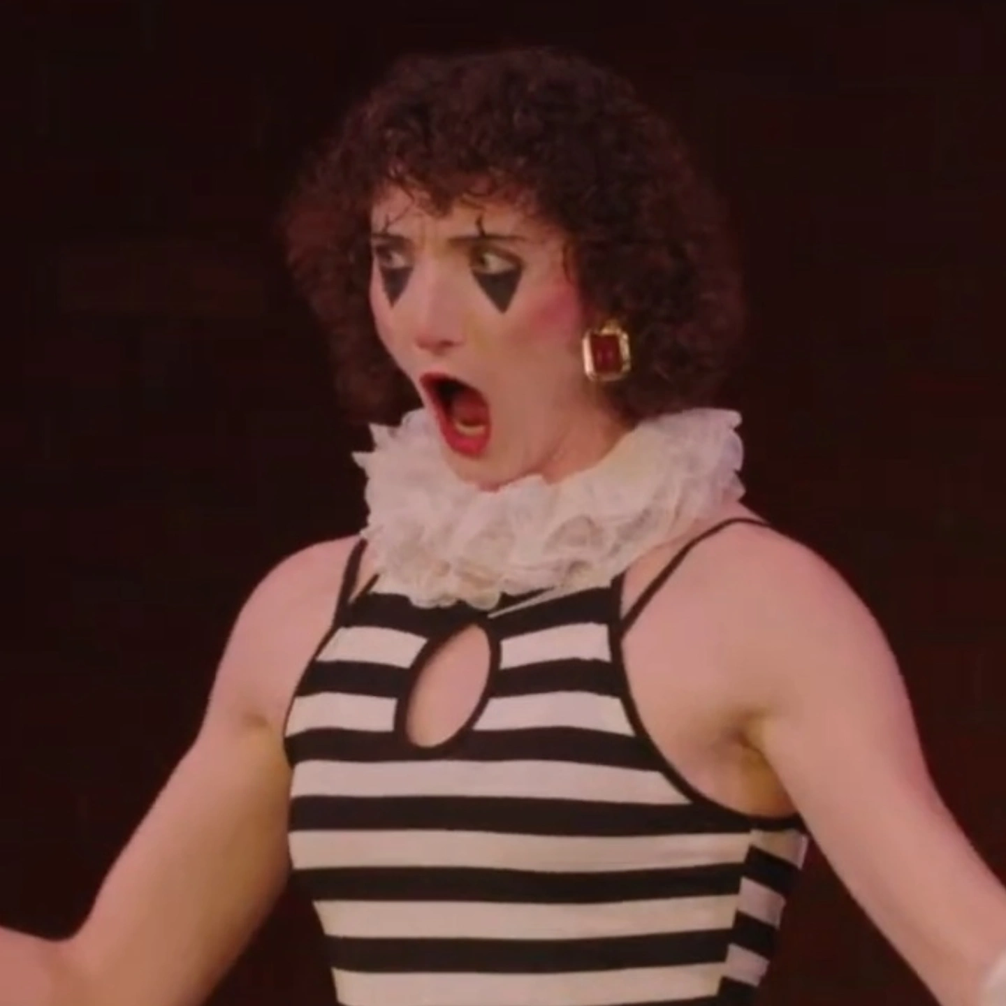 Person with curly hair, wearing striped top and ruffled collar, with surprised expression, open mouth, and earrings.