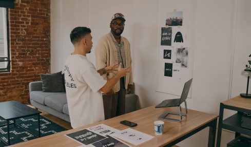 Two men talking in a modern office with a desk, laptop, and wall photos, one gesturing with hands, the other listening.