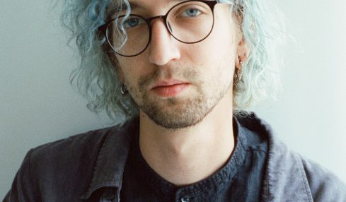 Young man with curly, light blue hair, glasses, and a serious expression, wearing a dark shirt and jacket, against a plain background.
