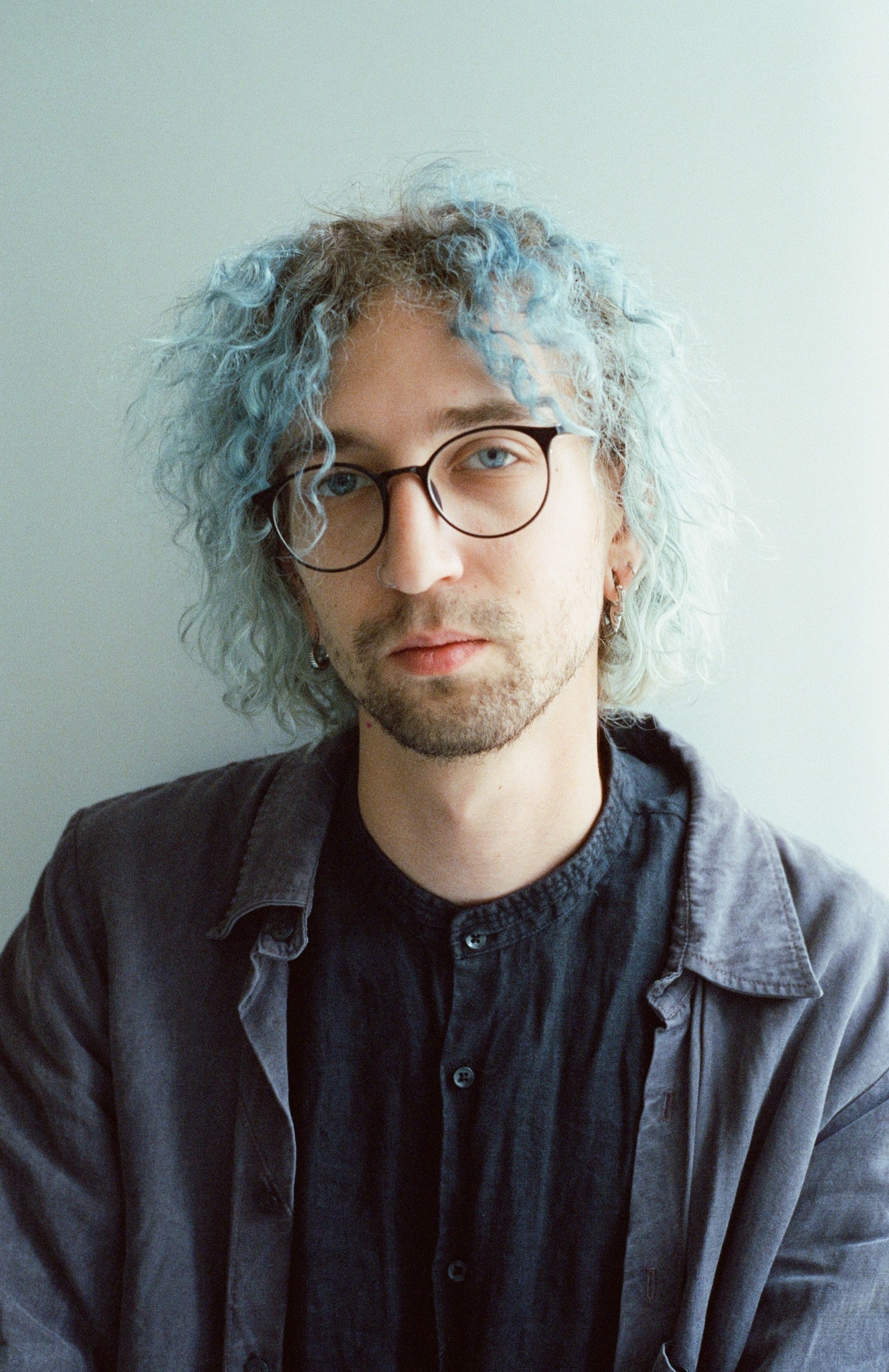 Young man with curly, light blue hair, glasses, and a serious expression, wearing a dark shirt and jacket, against a plain background.