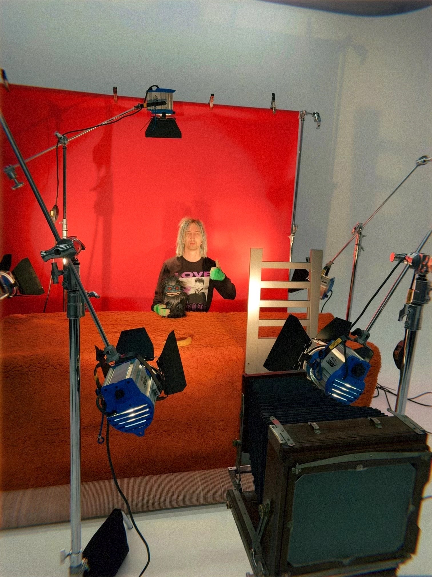 Person sitting at a desk in a studio with a red backdrop, surrounded by lighting and camera equipment.