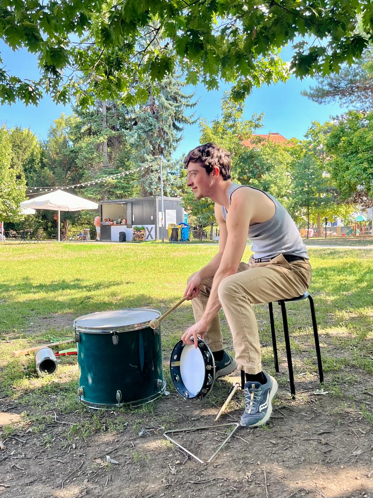 Young man sitting on a stool outdoors, playing drums with a drum set on grass under trees, sunny day.