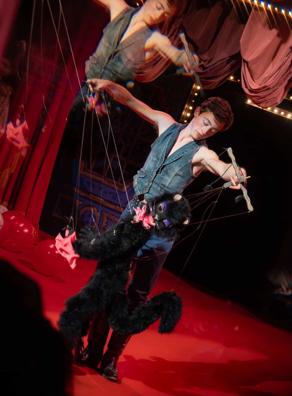 Circus performer in costume controls a black dog on a leash during a show, with red curtains and stage lighting in background.