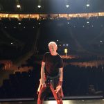 Person with light-colored hair standing on stage in an empty theater, facing away from the camera, with rows of seats behind.