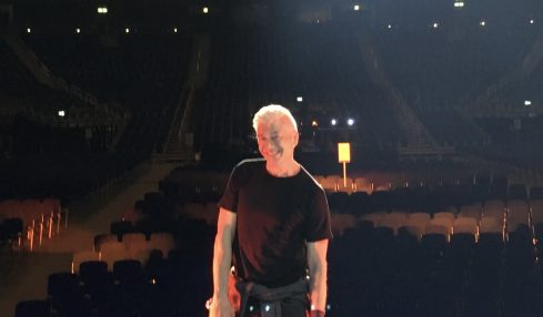 Person with light-colored hair standing on stage in an empty theater, facing away from the camera, with rows of seats behind.