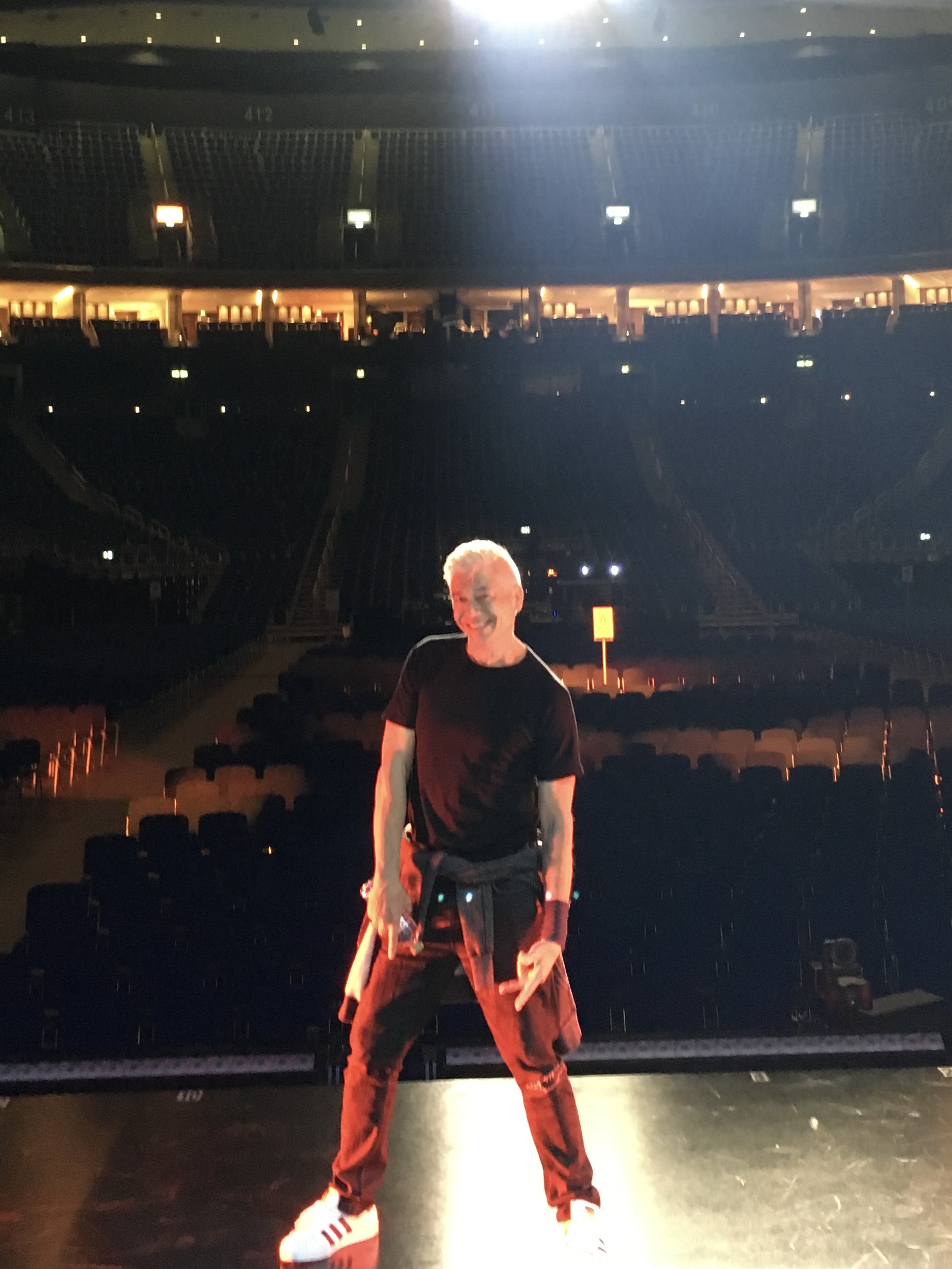 Person with light-colored hair standing on stage in an empty theater, facing away from the camera, with rows of seats behind.