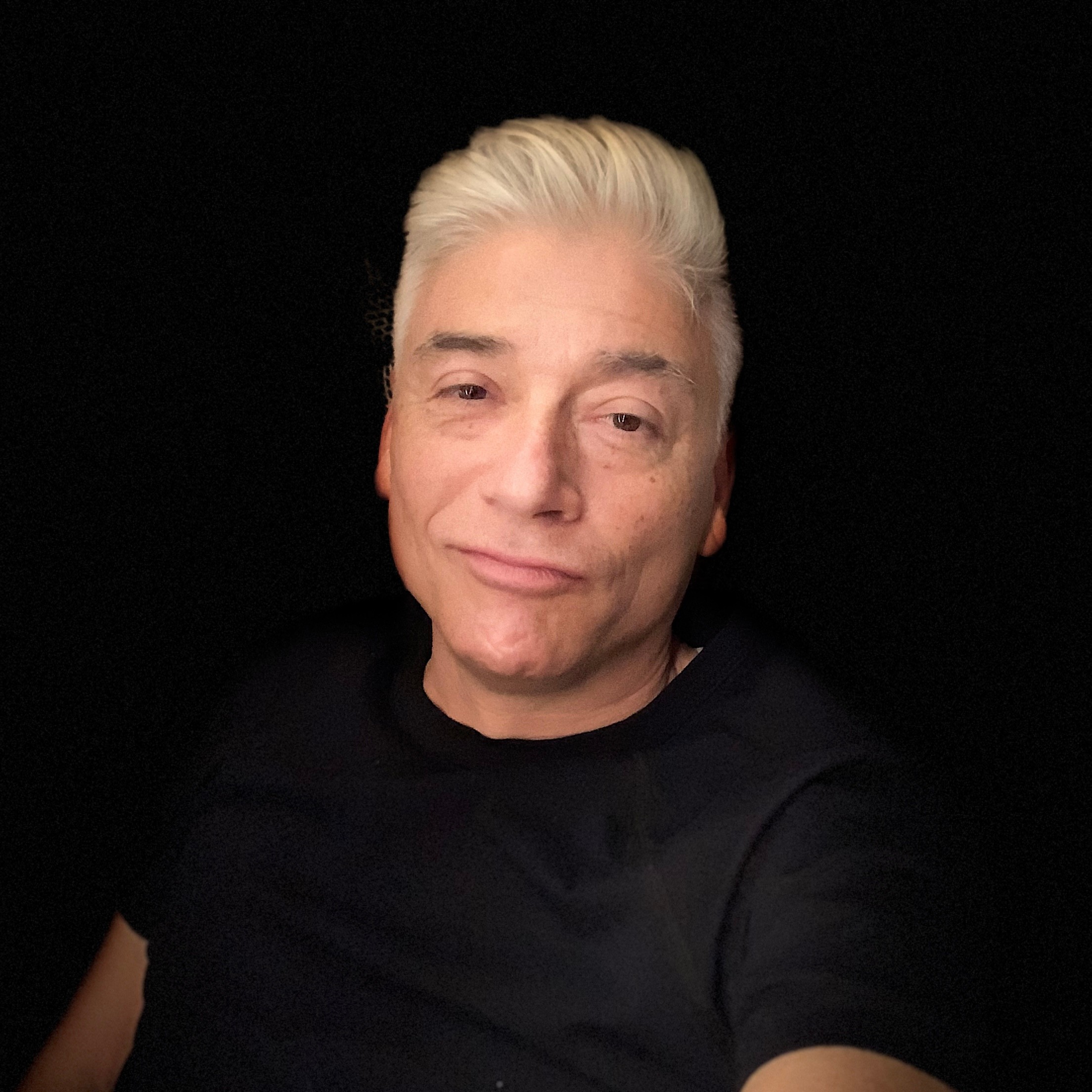 Person with short, platinum blonde hair and a slight smile, wearing a black shirt, against a dark background.