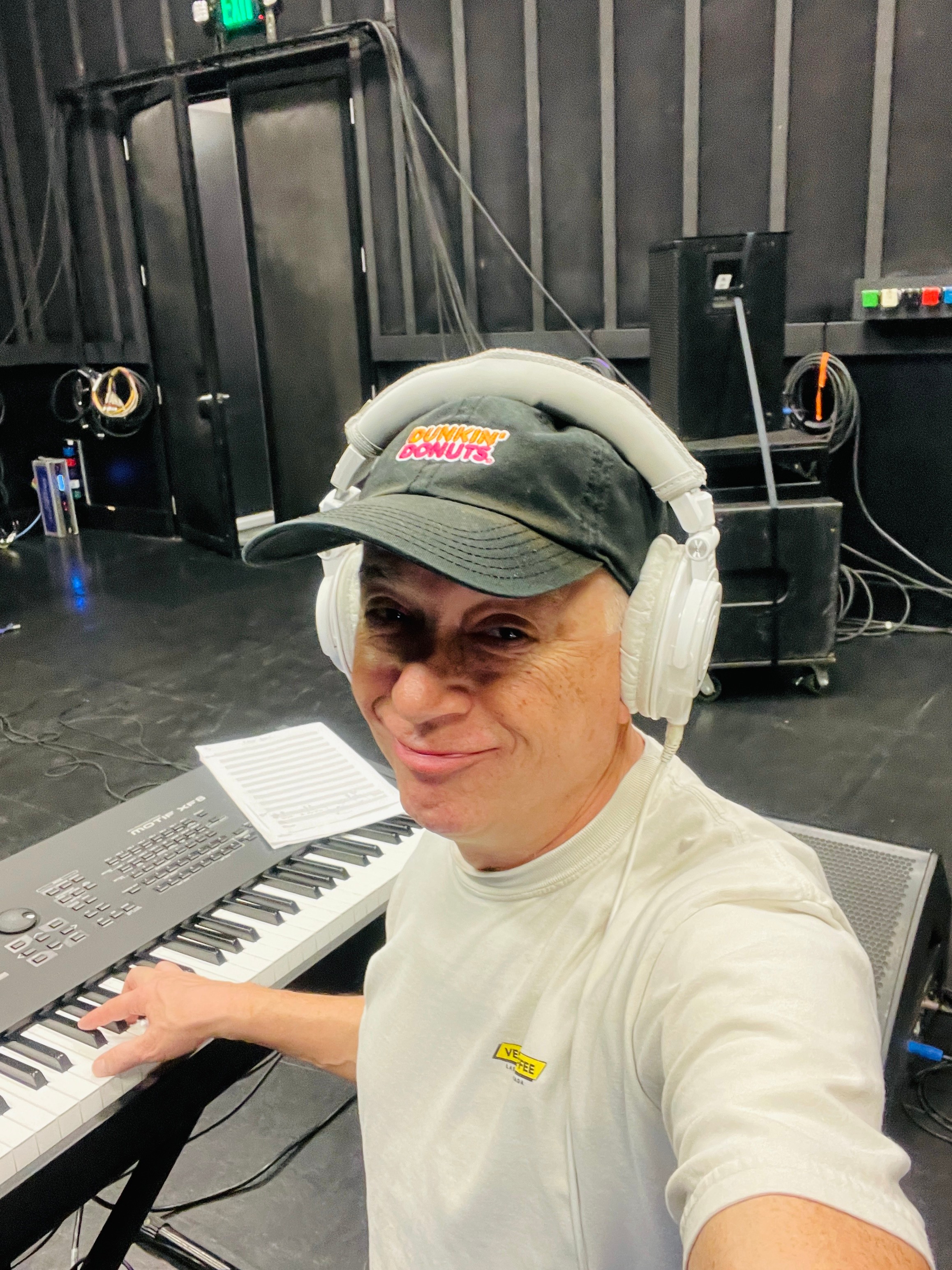 Person wearing headphones and a cap, smiling, sitting at a keyboard in a studio or stage setting.