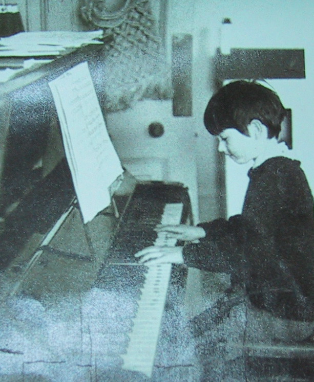 Child playing piano with sheet music, sitting upright, focused on keys, in a room with furniture and objects in background.