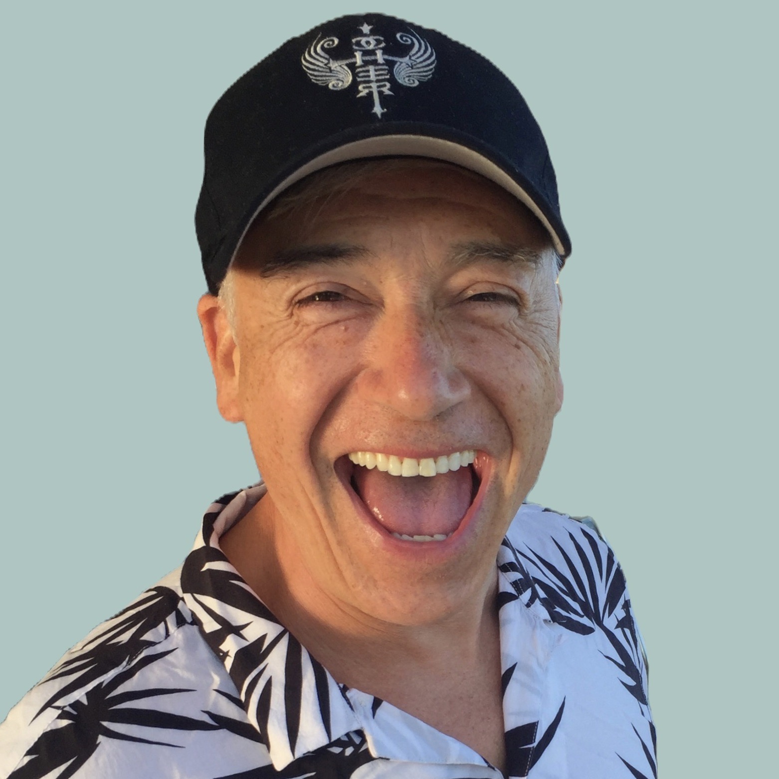 Person smiling and laughing, wearing a black cap and a patterned shirt, against a light blue background.
