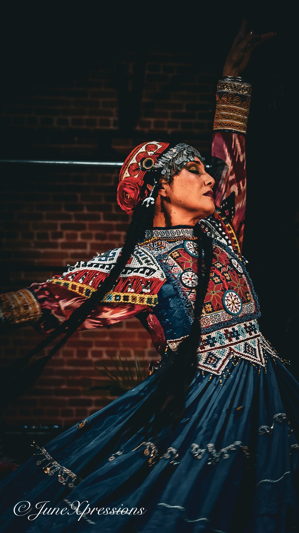 Woman in traditional attire dancing with one arm raised, wearing a decorated headpiece and colorful dress, brick wall background.