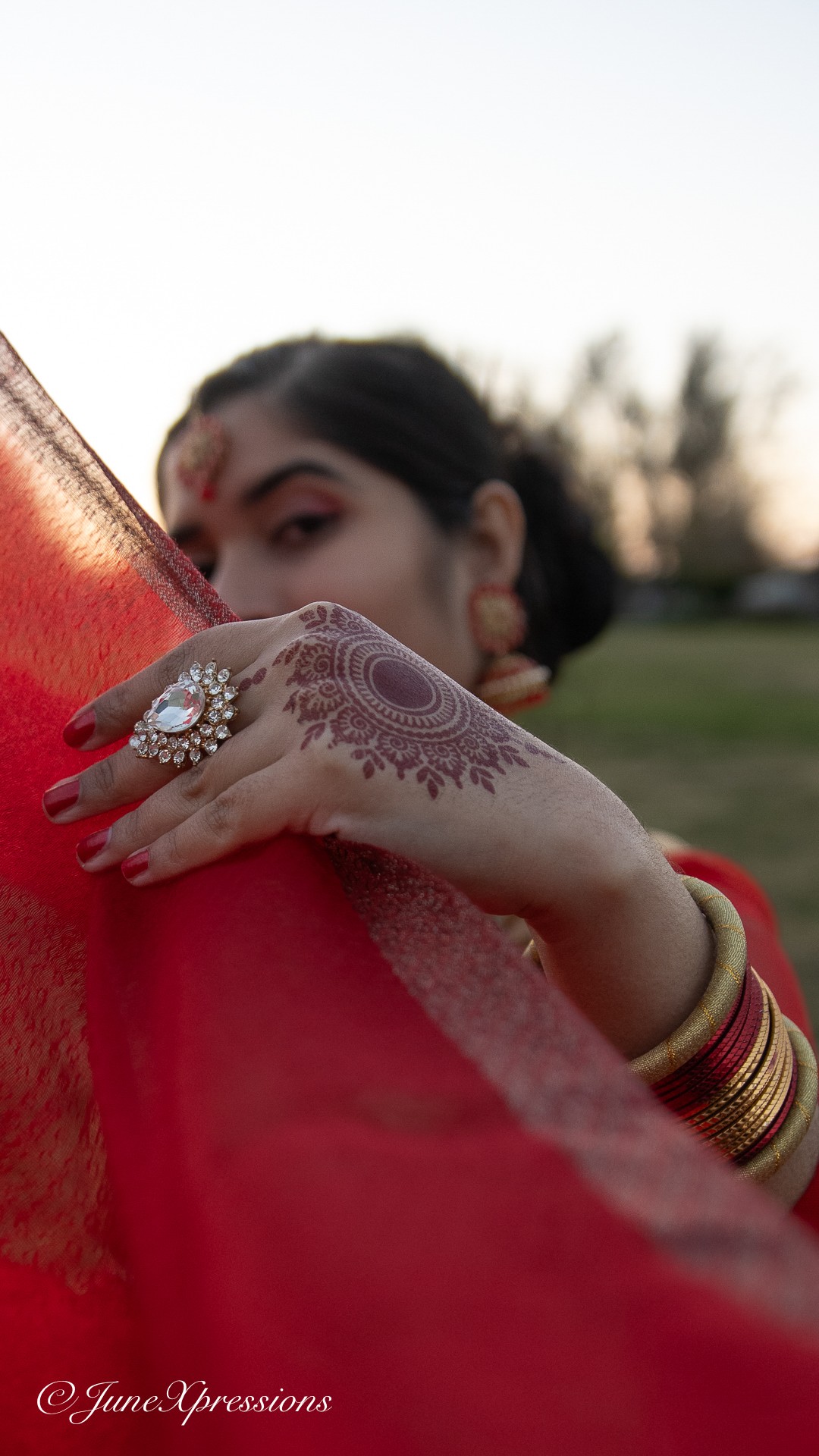 Person with henna on hand, wearing jewelry, holding red fabric outdoors, face partially visible, background blurred.