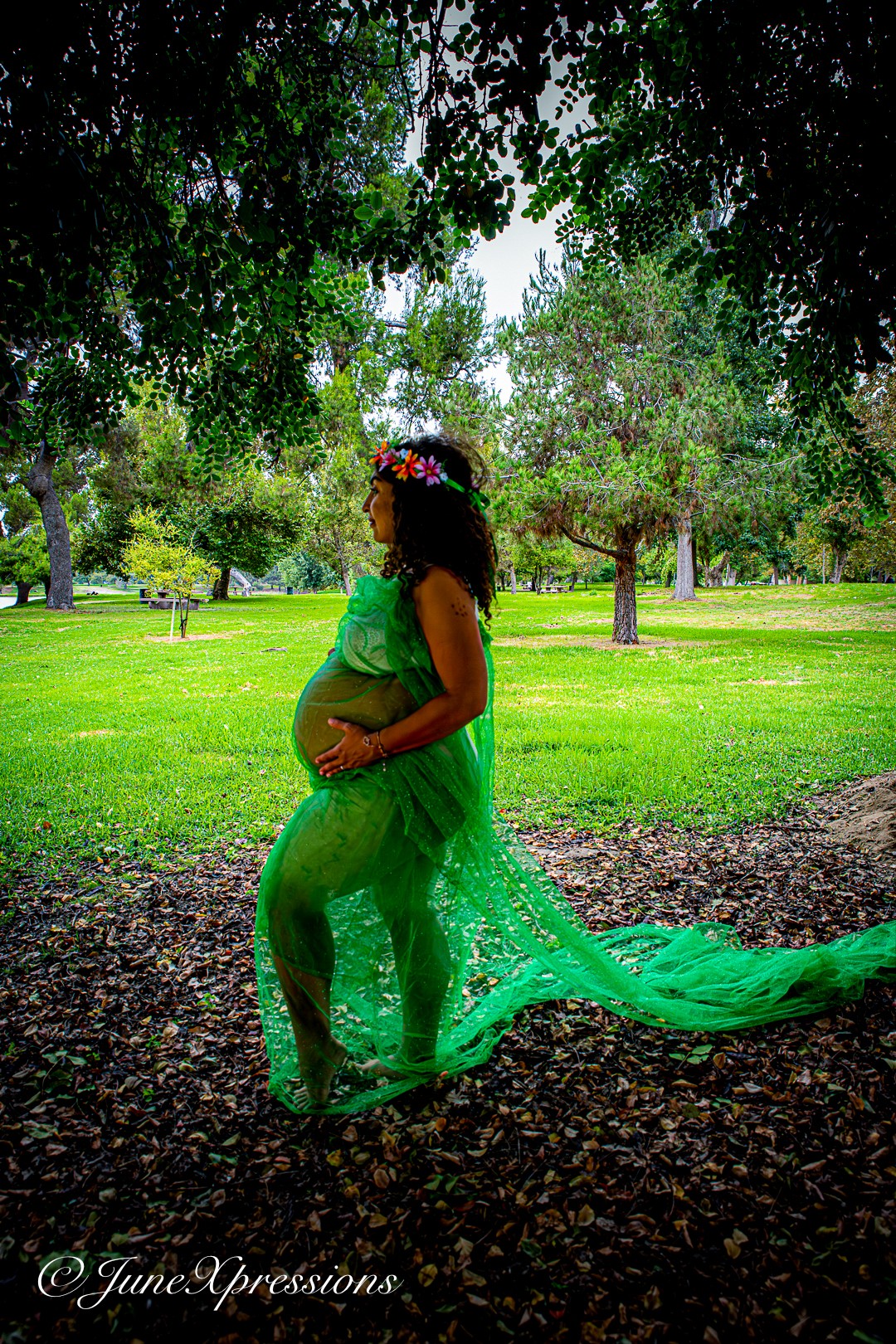 Pregnant woman in a green dress and floral headband standing outdoors in a park, holding her belly, surrounded by trees and grass.
