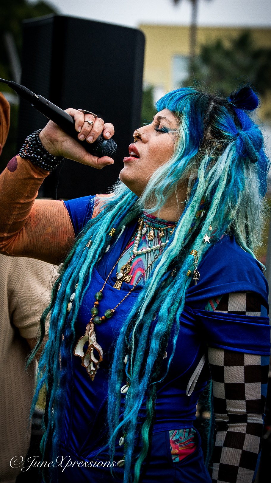 Person with blue and turquoise dreadlocks singing into a microphone outdoors, wearing colorful clothing and accessories.