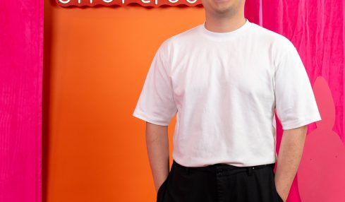 Young man with glasses in white t-shirt and black pants standing in front of colorful backdrop with text.