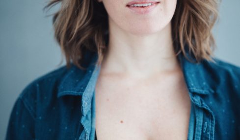Portrait of a woman with shoulder-length wavy brown hair, wearing a denim shirt over a black top, against a plain background.