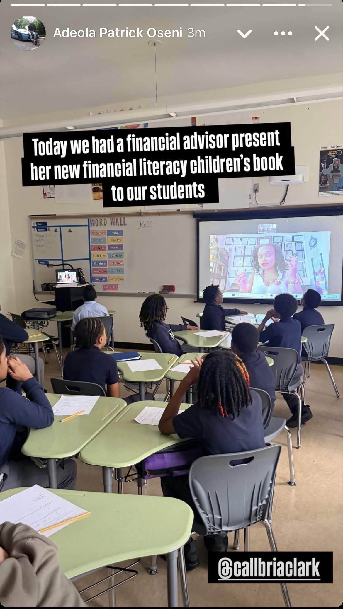Classroom with students watching a presentation on a large screen, teacher at the front, educational posters on wall.