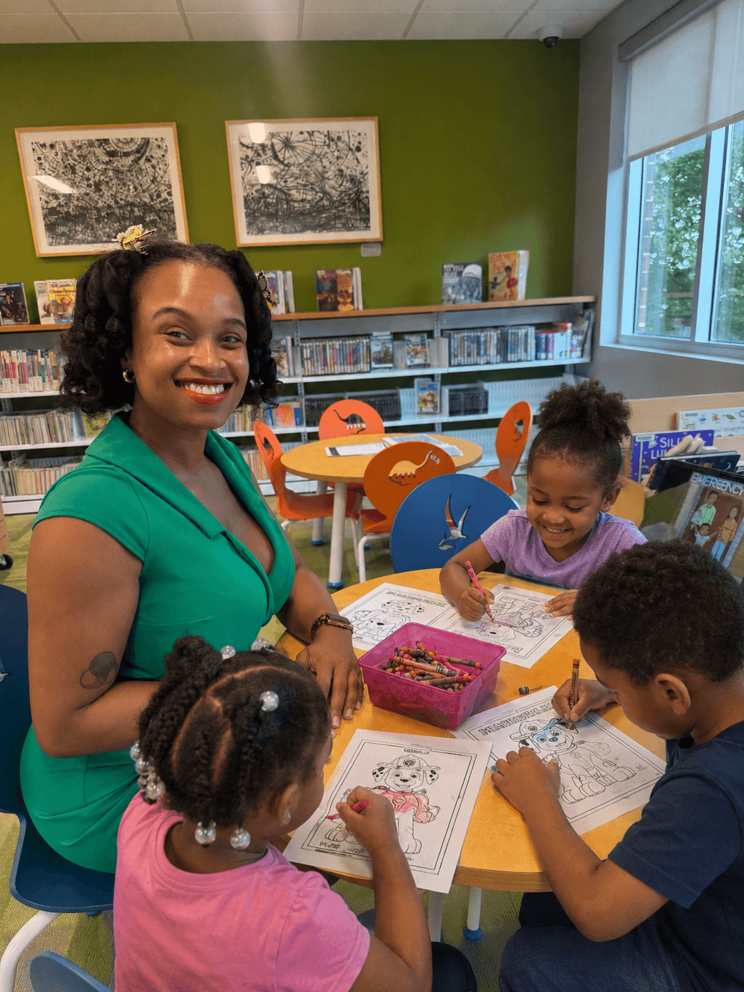 Smiling woman with children at a table in a library, coloring and reading books, green wall with framed pictures in background.