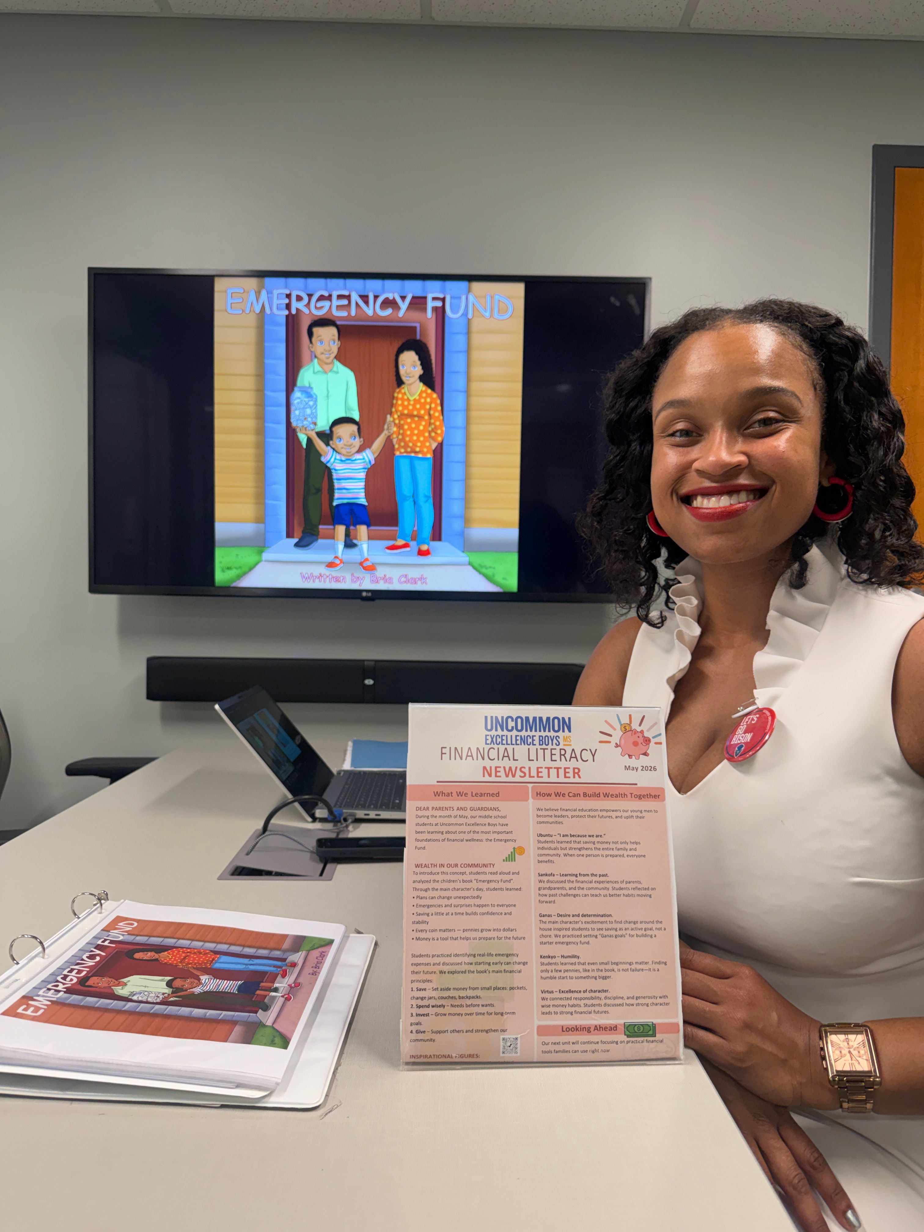 Two women sit at a table with a TV screen displaying a cartoon scene, and papers and a tablet on the table.