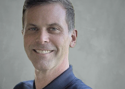 Portrait of a smiling man with short dark hair, wearing a dark collared shirt, against a plain background.
